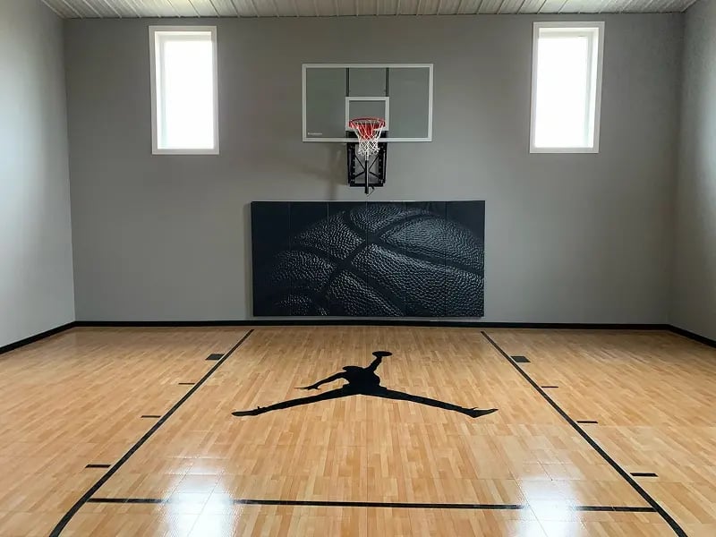 Custom indoor basketball court with hardwood floor, wall-mounted hoop, and padded wall panel built by Titus Contracting