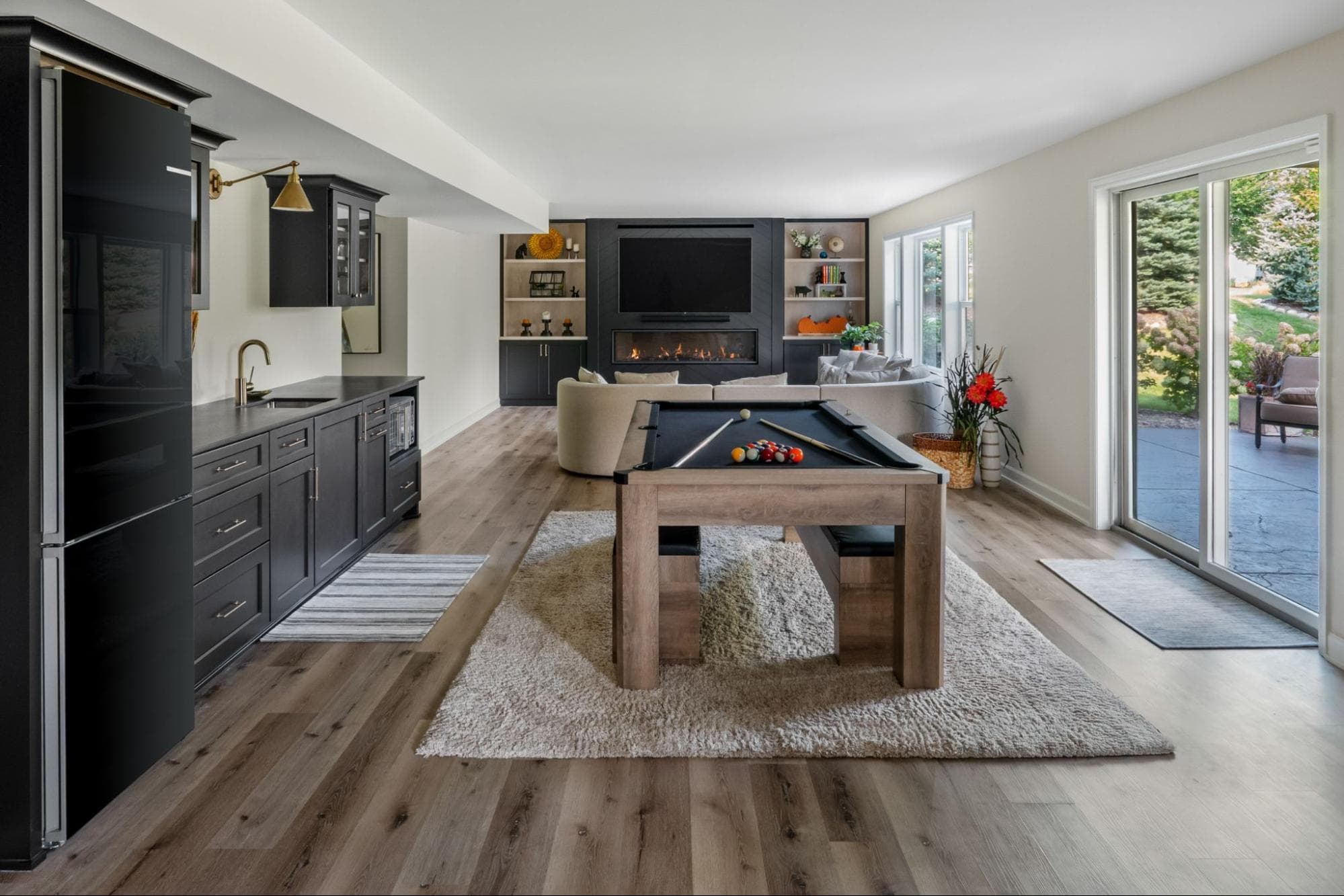 a wide shot of a finished basement, showcasing a full fridge, custom bar, pool table, and tv area with fireplace and built-in shelving.