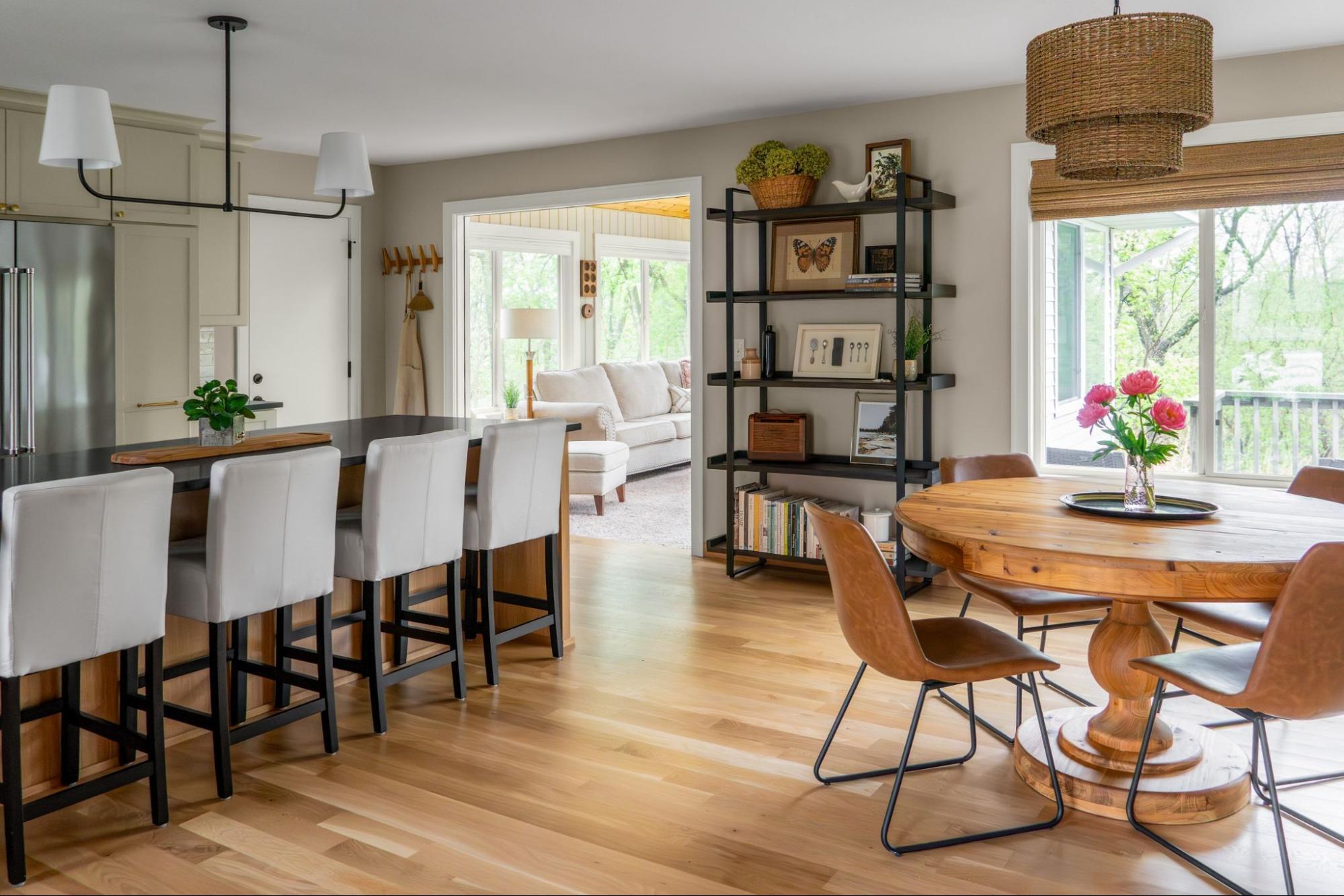 a dining area and kitchen with eclectic, yet cohesive design. a boho-inspired rattan chandelier in the dining room over a rough-hewn wooden table. an open industrial pipe bookshelf. Light green cabinets in the kitchen.