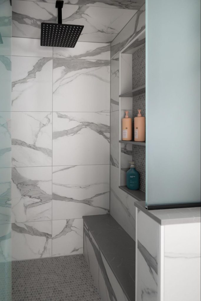 view inside a shower with a bench. the shower is lined with large-scale white marbled tiles. interior shelves above the bench offer storage.