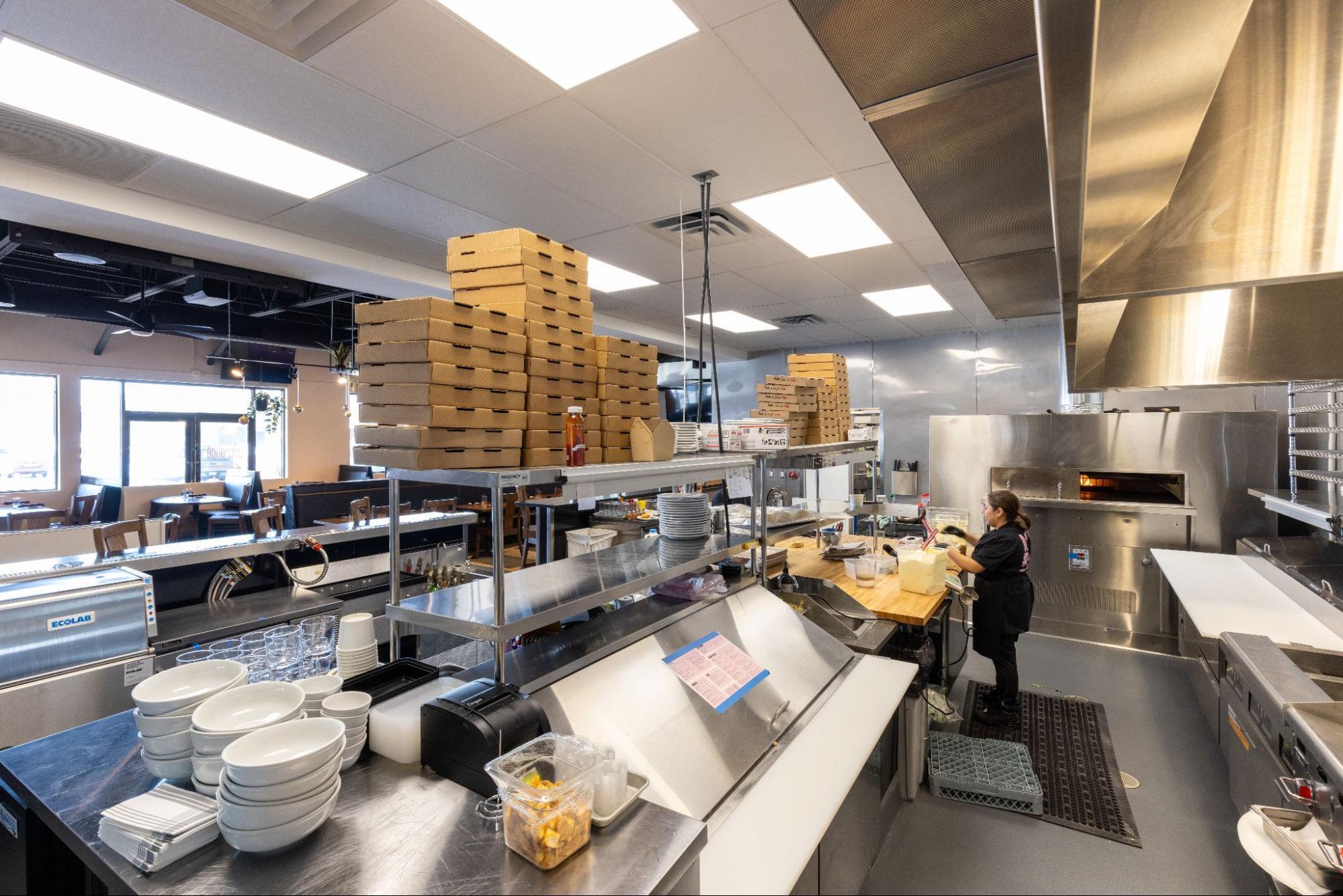 Commercial kitchen with prep station, stacked pizza boxes, and wood-fired oven in use