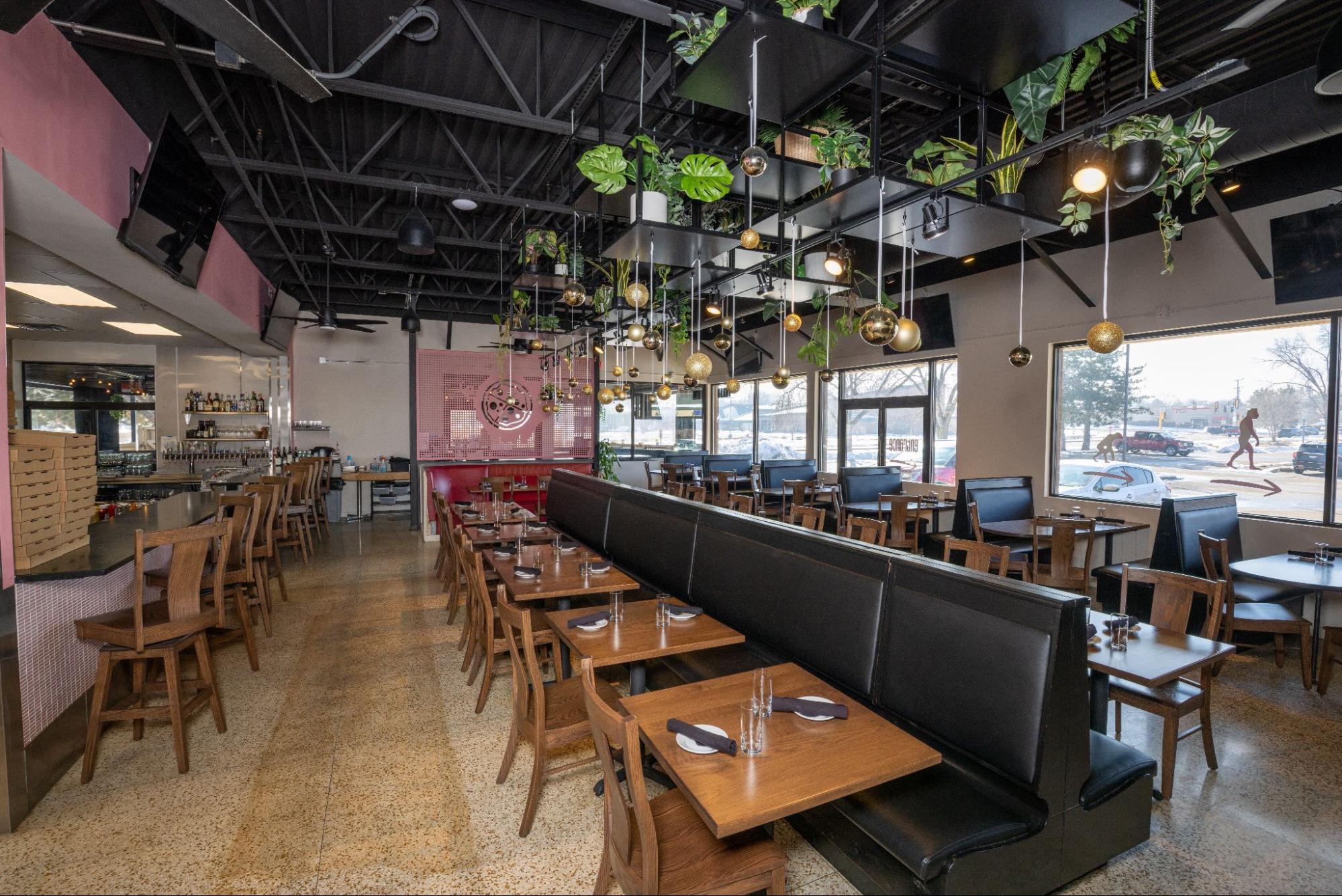 Modern restaurant dining area with booth seating, bar, large windows, and hanging plants and pendant lights
