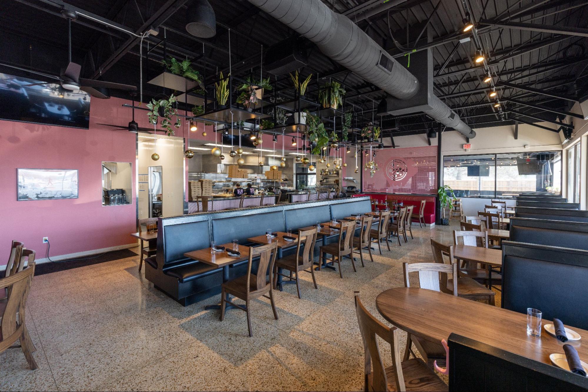 Spacious restaurant dining area with booth seating, open kitchen, and hanging plants with decorative lighting