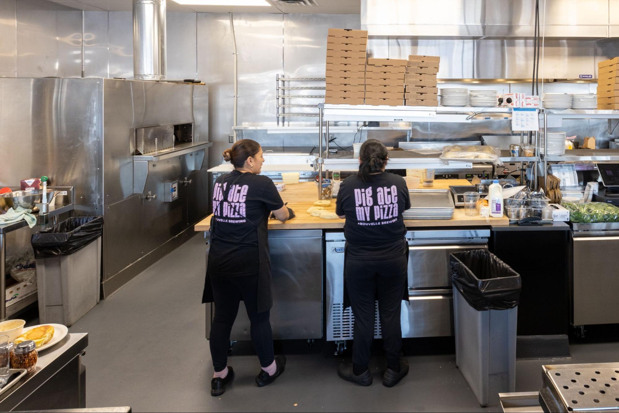 Restaurant kitchen staff preparing pizza dough at a stainless steel prep station with oven and stacked boxes