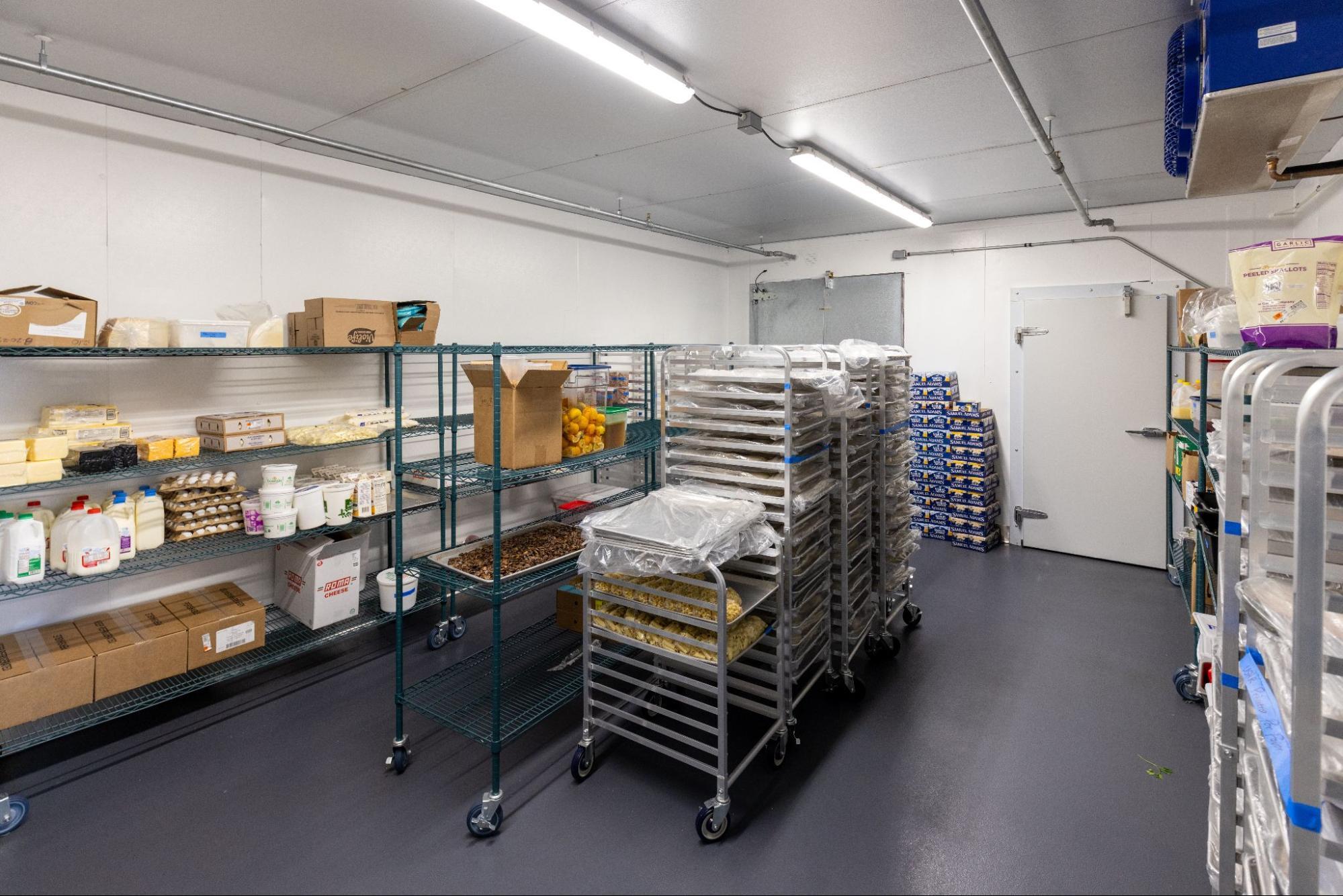 Commercial kitchen storage room with shelves of sauces, ingredients, and rolling racks of prepared food